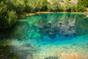 Eine tief türkisfarbene Karstquelle mit kristallklarem Wasser, in dem sich die grüne Vegetation und die felsigen Klippen spiegeln