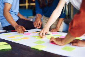Mehrere Personen stehen gemeinsam um einen Tisch und arbeiten kollaborativ an einer Aufgabe. Auf der Tischoberfläche liegen zahlreiche bunte Haftnotizen (in Gelb, Grün und Pink), die verteilt und teilweise beschrieben sind. Einige der Personen zeigen mit den Händen auf bestimmte Notizen oder halten Stifte, als würden sie Ideen besprechen, sortieren oder planen. Der Fokus liegt auf den Händen und der Interaktion, während die Personen selbst nur teilweise sichtbar sind. Die Szene vermittelt Teamarbeit, Brainstorming und kreative Zusammenarbeit in einer Arbeits- oder Workshop-Umgebung.